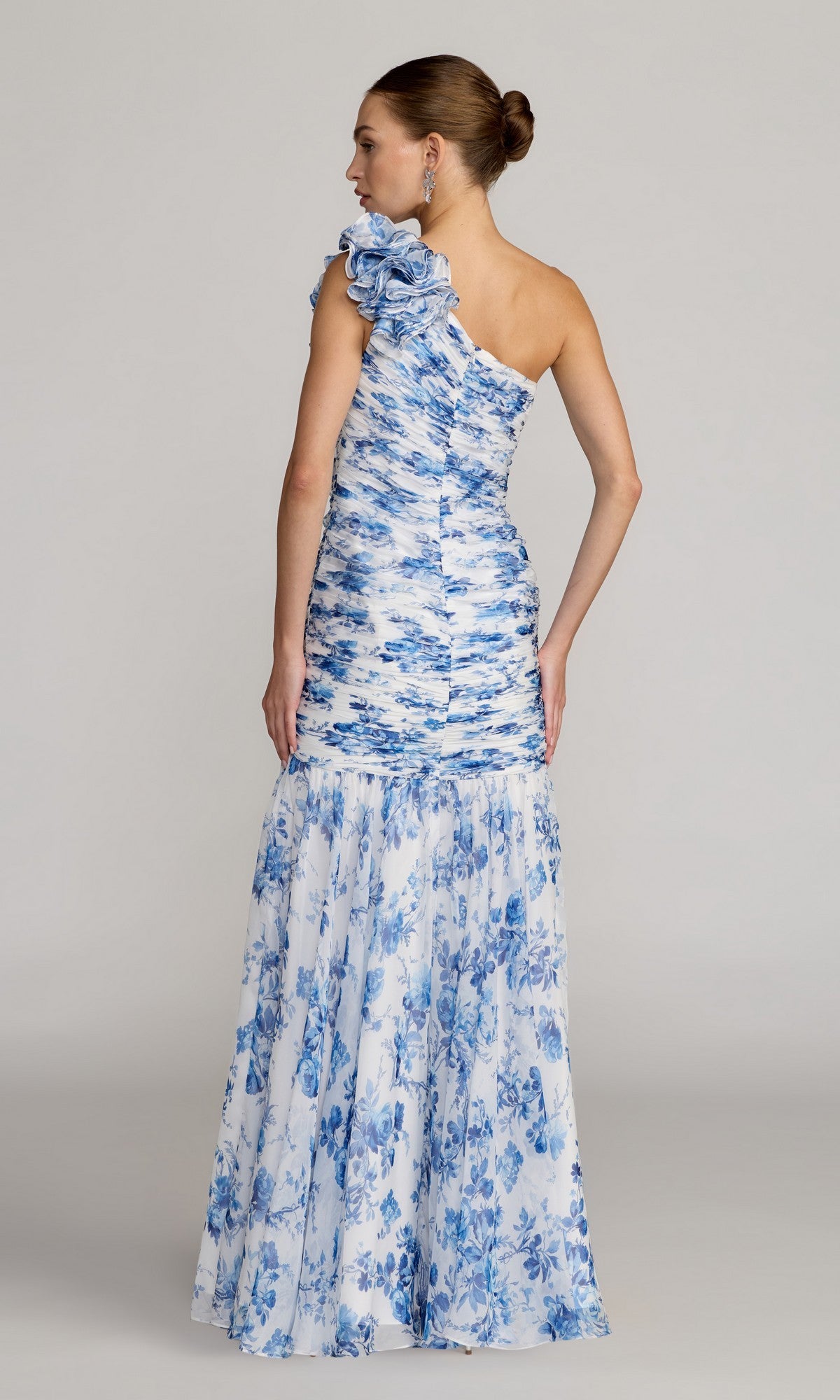 A woman stands with her back to the camera, wearing the Long Formal Dress: Mac Duggal 68992—a blue and white floral one-shoulder gown featuring a fitted bodice and flared skirt—set against a plain light background. This sophisticated ensemble captures the signature elegance of designer Mac Duggal.